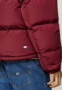 Jachetă puffer bordeaux cu un model în grilaj, având mansete ribbate și un patch cu logo-ul Tommy Jeans. Asortată cu blugi din bumbac albastri.