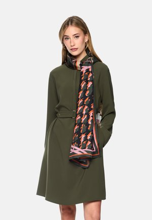 Woman wearing an olive green belted dress and a black, orange, and pink patterned scarf around her neck, standing with one hand in pocket.