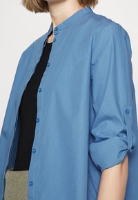 Ljusblå skjorta med ståkrage, uppkavlade ärmar och svart underlager. Tillverkad av ett slätt tyg med subtil lyster.