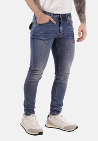 Uomo che indossa jeans slim blu e sneakers bianche, con una mano nella tasca e una manica tatuata visibile sul braccio destro.