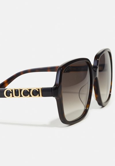 Brýlové sluneční brýle v želvovinovém designu s masivním rámem, gradientní šedé čočky a zlatým nápisem "GUCCI" na rámu. Hladký plastový povrch.