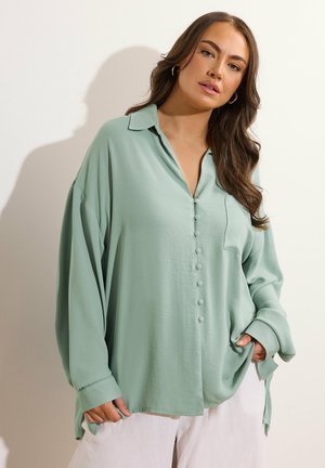 Vrouw met lang bruin haar draagt een lichtgroene oversized blouse met knopen en witte broek, poserend tegen een lichte achtergrond.