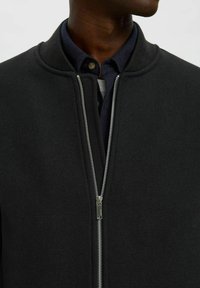 Giacca nera con zip realizzata in tessuto liscio, con colletto a costine, zip metallica e un look a strati con una maglietta scura sotto.
