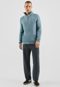 Jasnoniebieski sweter z suwakiem pod szyją wykonany z teksturowanej tkaniny, zestawiony z ciemnoszarymi dżinsami i czarnymi butami z białymi akcentami.