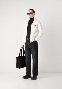 Felpa bianca con zip e logo rosso, dolcevita nero, jeans neri, scarpe nere e borsa a tracolla nera. Il modello è in piedi davanti a uno sfondo semplice.