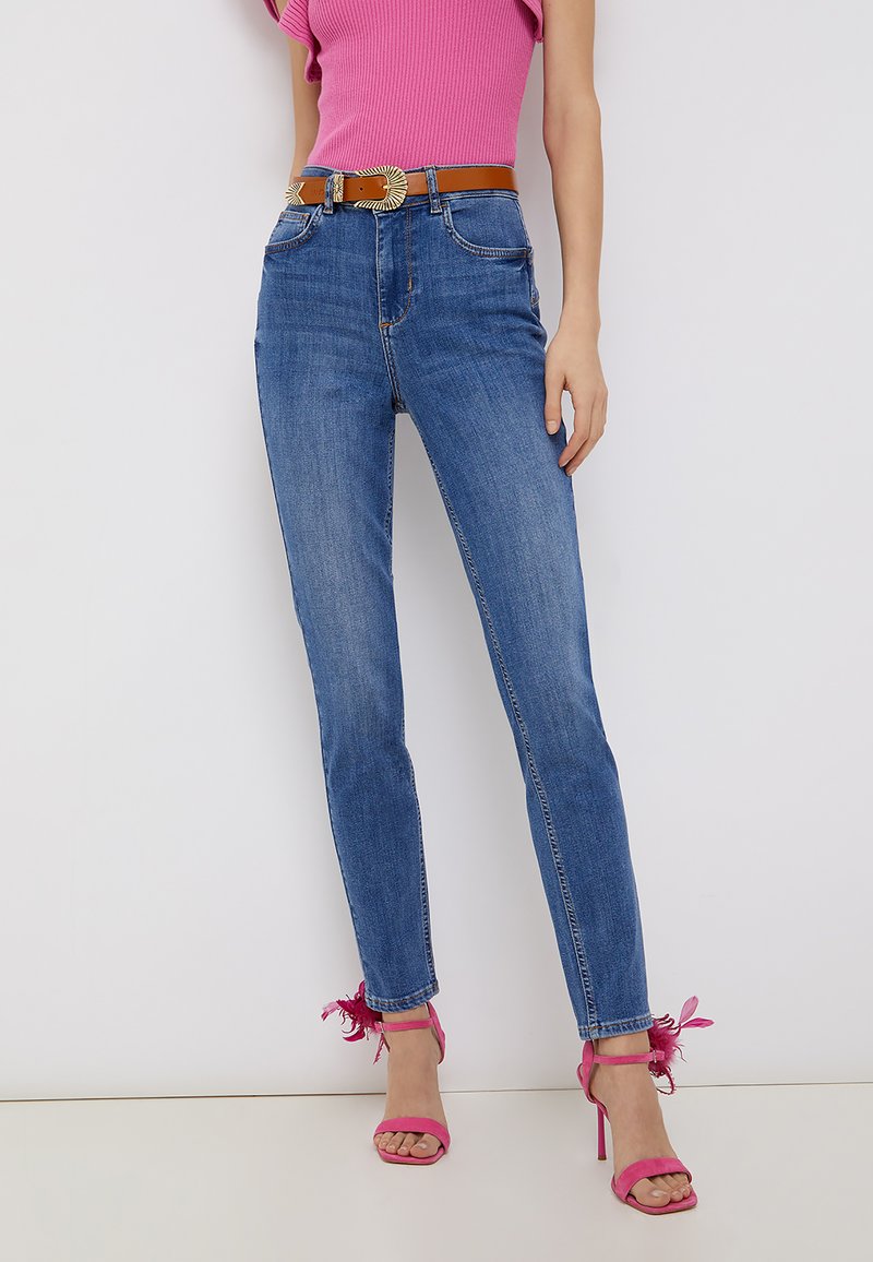 Jeans skinny blu in denim, abbinati a una maglia rosa a costine. Gli accessori includono una cintura marrone con fibbia dettagliata e tacchi alti rosa.