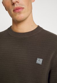 Pulover tricotat maro cu o textură ribbed, guler rotund și un mic eticheta logo pe piept. Afișat într-un format de prim-plan.