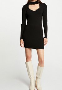 Robe en tricot côtelé noir à manches longues, décolleté en V profond et détail de choker. Portée avec des bottes blanches hautes, mettant en avant des couleurs et des textures contrastées.
