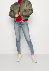 Chaqueta bomber oliva con puños acanalados, suéter de punto rojo, jeans ajustados de color azul claro y zapatillas grises con diseño texturizado y cordones.