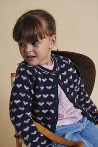 Granatowy kardigan w różowe i białe wzory serc, z okrągłym dekoltem i drewnianymi guzikami, noszony na kraciastą koszulę i dżinsowe spodnie.