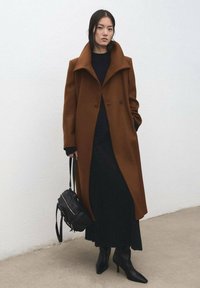 Bruine wollen jas met een brede kraag en twee knopen, gecombineerd met een zwarte trui en een lange zwarte rok. Zwarte puntige enkellaarsjes en een tas.