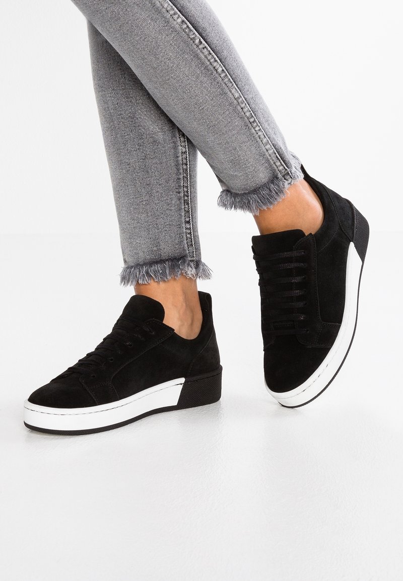 Jambes portant un jean gris à ourlet effiloché et des baskets en daim noir avec semelles plateformes blanches sur un sol blanc.