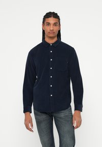 Camisa de pana azul marino con cuello, cierre de botones y un bolsillo en el pecho. Mangas largas con puños abotonados, combinada con vaqueros oscuros.
