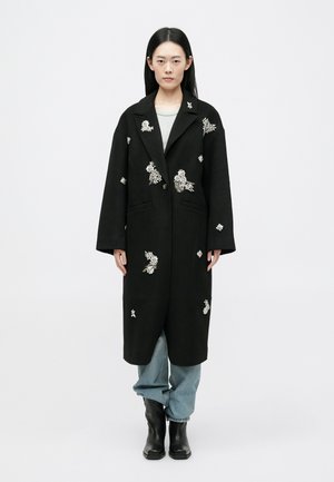 Juodas ilgas paltas su gėlių siuvinėjimais, dideliais apykaklėmis, sagų uždarymu, šoninėmis kišenėmis, derinamas su šviesiais džinsiniais kelnais ir juodais kulkšnies batais.