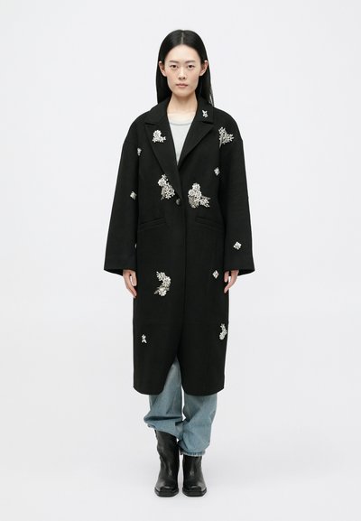 Cappotto lungo nero con ricami floreali, ampi risvolti, chiusura con bottoni, tasche laterali, abbinato a pantaloni di jeans chiari e stivaletti neri.