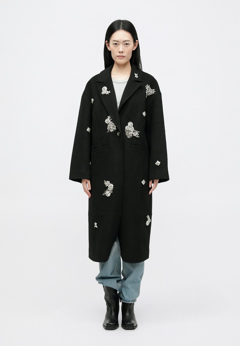 Črn dolg plašč z ročno vezenim cvetličnim vzorcem, širokimi reverji, gumbnim zapiranjem in stranskimi žepi, v kombinaciji s svetlimi denim hlačami in črnimi gležnjarji.