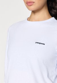 Bijela majica dugih rukava od mekog pamuka, s crnim "Patagonia" logom na lijevoj strani grudi i klasičnim dekolteom.