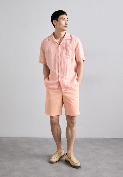 Camicia in lino rosa chiaro con maniche corte e taschino sul petto, abbinata a pantaloni corti pesco e scarpe slip-on beige con dettaglio intrecciato.