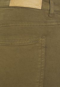 Calças de ganga verde-azeitona com um retalho de pele bege. Apresentam um corte reto, costura visível e um detalhe de bolso lateral.