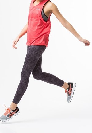 Rotes ärmelloses Tanktop über schwarzer gemusterter Leggings, kombiniert mit grau-orangefarbene Sportschuhen, Modell in Bewegung vor einem schlichten Hintergrund.