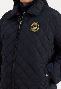 Quiltet svart jakke med høy krage, med en gull- og rød brodert logo og en sidelommer med knappelukking.
