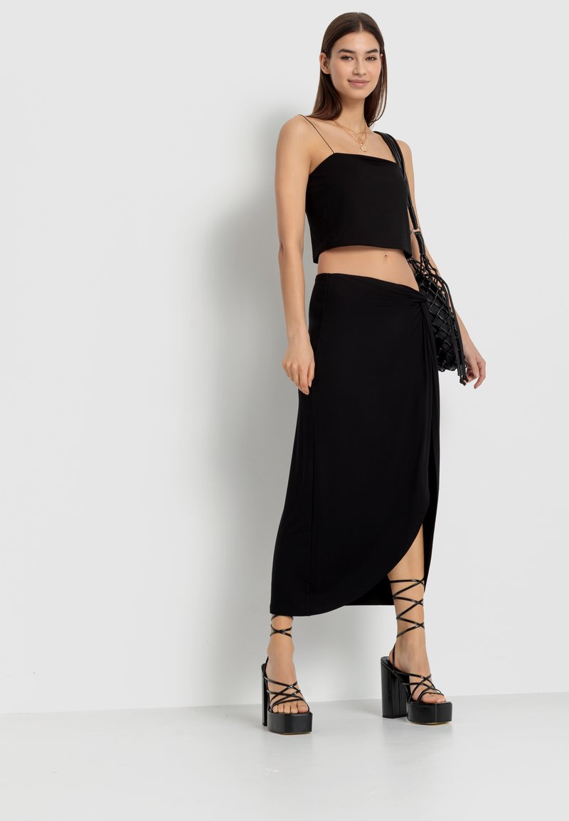 Schwarzes Cropped Top mit Spaghetti-Trägern, kombiniert mit einem langen schwarzen Rock mit hohem Schlitz. Das Schuhwerk hat dicke Plateausohlen und überkreuzte Riemen.