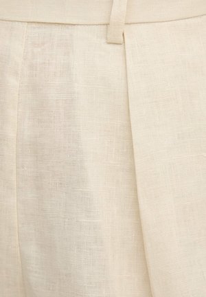 Primo piano di pantaloni in lino beige che mostrano il tessuto strutturato, una passante per cintura e un dettaglio di piega frontale.