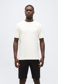 Άνδρας που στέκεται αντιμετωπίζοντας μπροστά, φοράει απλό λευκό κοντομάνικο μπλουζάκι με μικρό κείμενο και μαύρα σορτς, σε ουδέτερο φόντο.