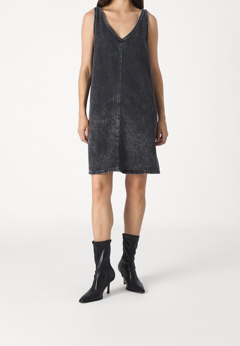 Femme portant une robe en jean noire délavée sans manches avec un col en V et des bottes noires à talons mi-mollet, debout sur un fond clair uni.