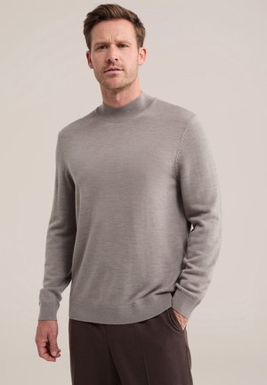 Homme portant un pull gris clair à manches longues avec col montant et un pantalon marron foncé, debout devant un fond beige uni.