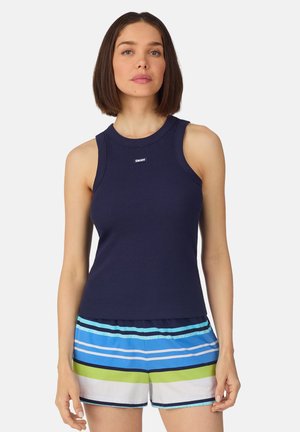 Vrouw met steil bruin haar, gekleed in een marineblauw mouwloos topje en gestreepte blauwe, groene en witte korte broek, staande tegen een effen achtergrond.