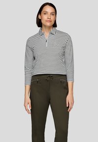 Gestreiftes Langarm-Poloshirt in Schwarz und Weiß mit halbem Reißverschluss am Kragen und einer kleinen Tasche mit dekorativen Akzenten. Kombiniert mit olivgrünen Hosen.