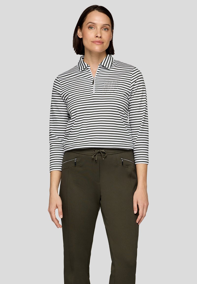 Gestreiftes Langarm-Poloshirt in Schwarz und Weiß mit halbem Reißverschluss am Kragen und einer kleinen Tasche mit dekorativen Akzenten. Kombiniert mit olivgrünen Hosen.