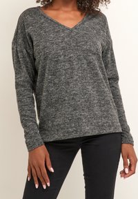 Pull gris à manches longues avec col en V et finition texturée. Le tissu semble doux et légèrement duveteux, avec une coupe décontractée. Associé à un pantalon noir.