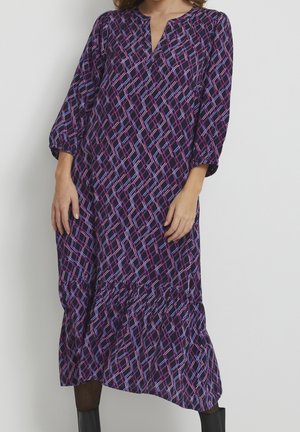 Kvinde iført en løs, lang lilla kjole med pink og blå geometriske linjer, trekvartærmer og sorte ankelstøvler.