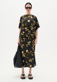 Top floreale nero con maniche corte, abbinato a una gonna floreale nera coordinata. Presenta un colletto arricciato, tiene una borsa in pelle nera e indossa sandali.