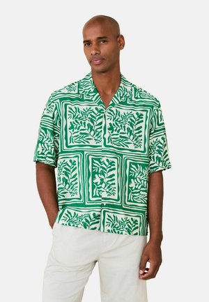 Mann trägt grünes und weißes kurzärmeliges Hemd mit Knöpfen und Blattmuster sowie hellfarbene Hose, steht mit einer Hand in der Tasche.