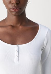 White ribbed long-sleeve top with a scoop neckline, featuring three metal snap buttons centered at the chest. Smooth texture.