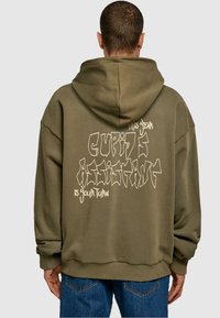 Olíva zöld, oversized kapucnis pulóver, hátul nagy grafikai dizájnnal, fehér szöveggel, amely tartalmazza a "Cupid's Assistant Is Your Turn" kifejezést.