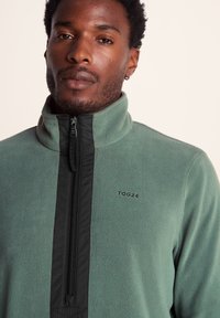 Fleece pullover with a high collar, featuring a zippered front and black accents. Main color is muted green with "TOG24" logo in black.