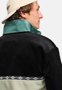 Men's corduroy jacket featuring a black body, teal collar, and textured green lower section. Includes a patterned black band at the waist.
