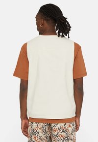 Gilet beige chiaro sopra una camicia a maniche corte marrone. Il gilet presenta un design pulito, mentre i pantaloni hanno una stampa leopardata in vari colori.