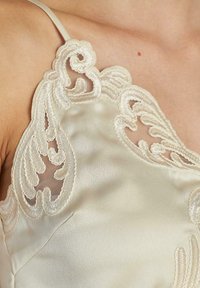 Zoom de un camisón de satén beige con intrincados bordados de encaje en el escote y tirantes finos sobre piel desnuda.
