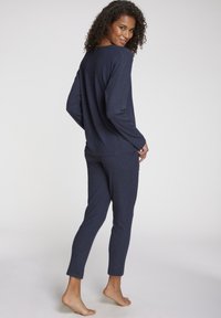 Woman standing barefoot, looking back over shoulder, wearing matching navy ribbed long-sleeve top and ankle-length pants against plain background.