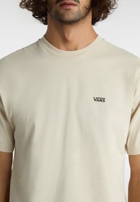 Muž v béžovém tričku Vans s malým černým logem Vans na levé části hrudi, zkráceném pod nosem a nad pasem.