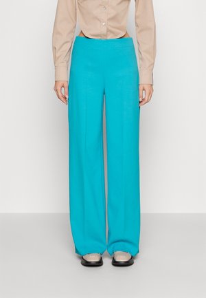 Persoon draagt hoge taille, wijde turkooizen broek, met een beige shirt erin gestopt en beige schoenen met zwarte zolen, staand tegen een witte achtergrond.