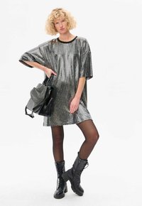 Robe métallisée argentée surdimensionnée avec des manches courtes, bord noir, portée avec des leggings noirs et des bottes de combat, tenant un sac à main noir.