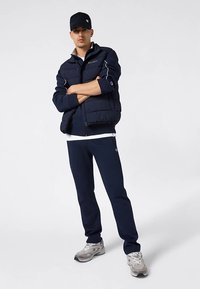 Modrá prešívaná bunda s bielymi detailmi, kombinovaná s modrými teplákmi. Šedé tenisky a zladená čiapka dopĺňajú športový vzhľad.