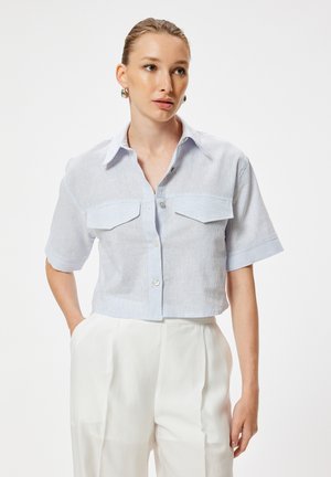 Vrouw met lichtblauw kortermig knoopoverhemd en witte broek met hoge taille, staand met één hand in de zak, kijkt iets naar rechts.