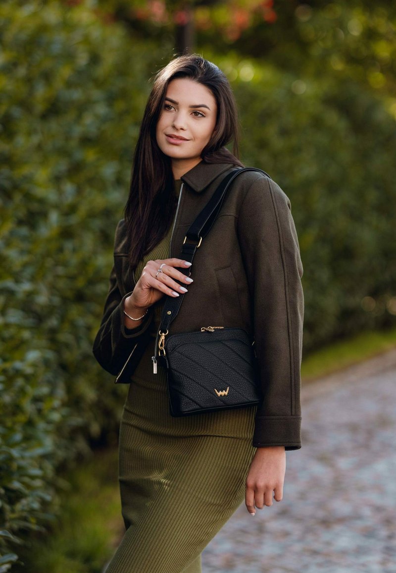 Černá koženková křížová taška s prošívaným designem, zlatými doplňky a nastavitelným popruhem; stylizováno s olivově zeleným outfitem a tmavou bundou.